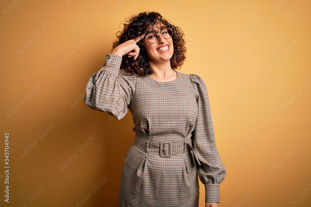 Beautiful arab business woman wearing dress and glasses standing over yellow background Smiling pointing to head with one finger, great idea or thought, good memory Beautiful arab business woman wearing dress and glasses standing over yellow background Smiling pointing to head with one finger, great idea or thought, good memory