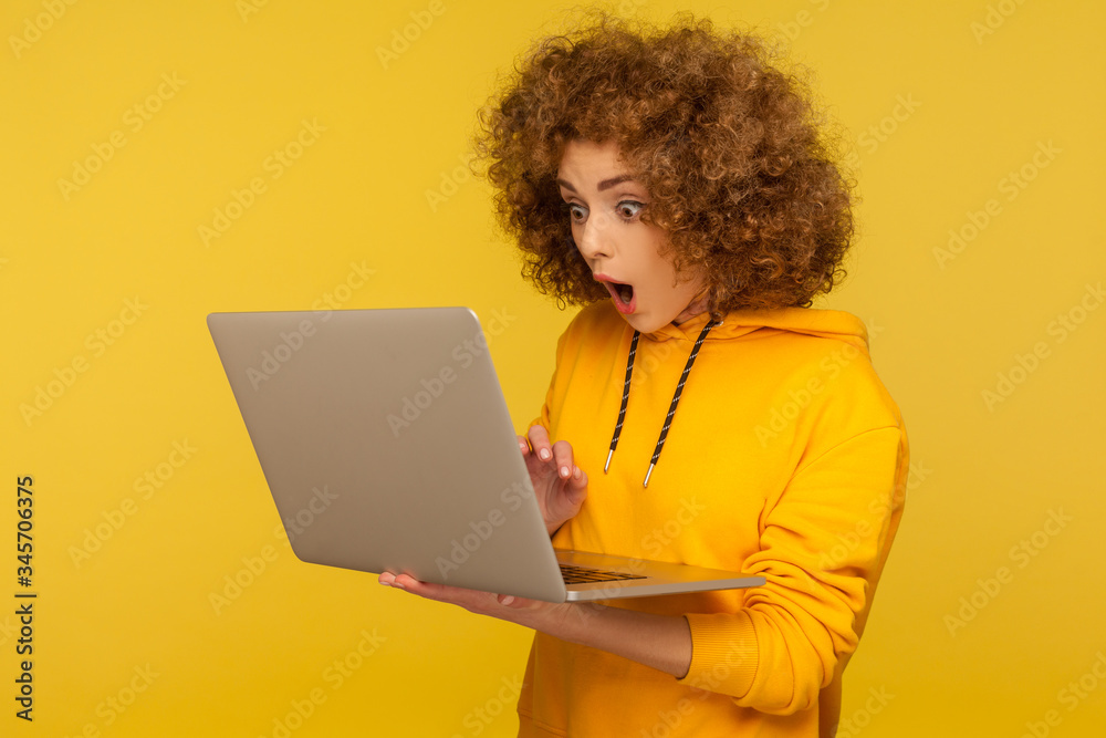 Surprised emotional bright woman with curly hair in urban style hoody looking at laptop screen with shocked expression, typing and browsing internet. indoor studio shot isolated on yellow background Surprised emotional bright woman with curly hair in urban style hoody looking at laptop screen with shocked expression, typing and browsing internet. indoor studio shot isolated on yellow background