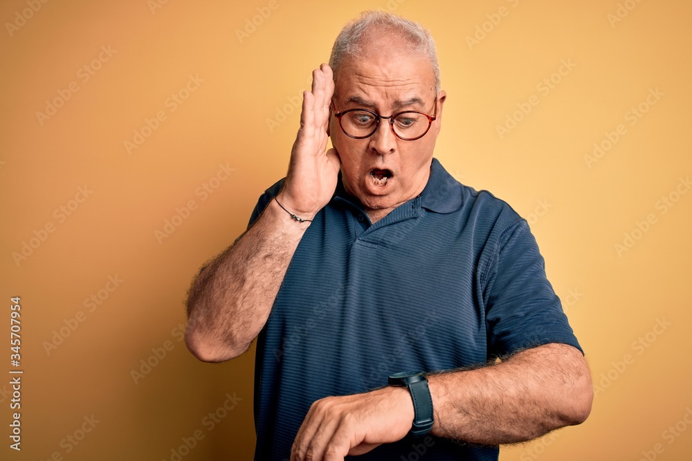 Middle age handsome hoary man wearing casual polo and glasses over yellow background Looking at the watch time worried, afraid of getting late Middle age handsome hoary man wearing casual polo and glasses over yellow background Looking at the watch time worried, afraid of getting late