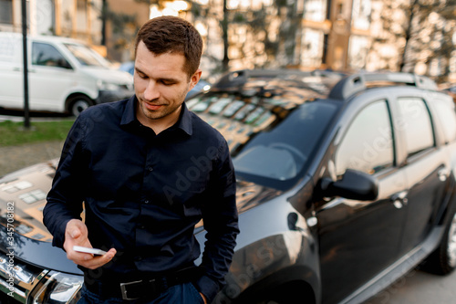 A young man or businessman in a dark shirt stands on the street near the car will look at the smartphone. The driver is waiting for his passenger or client. Urban transport concept