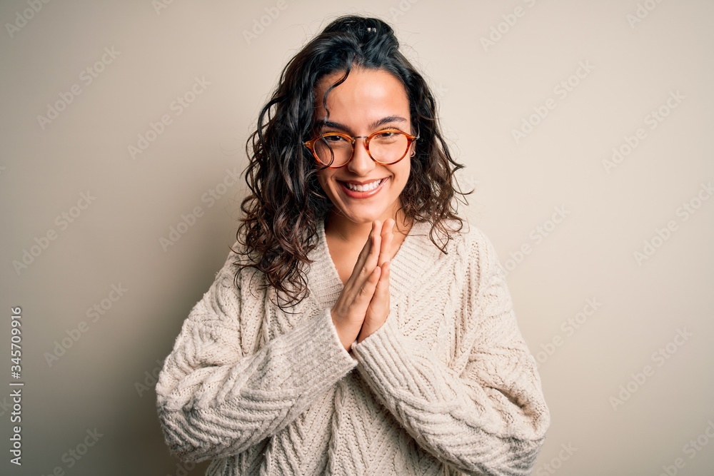 Beautiful woman with curly hair wearing casual sweater and glasses over white background praying with hands together asking for forgiveness smiling confident. Beautiful woman with curly hair wearing casual sweater and glasses over white background praying with hands together asking for forgiveness smiling confident.