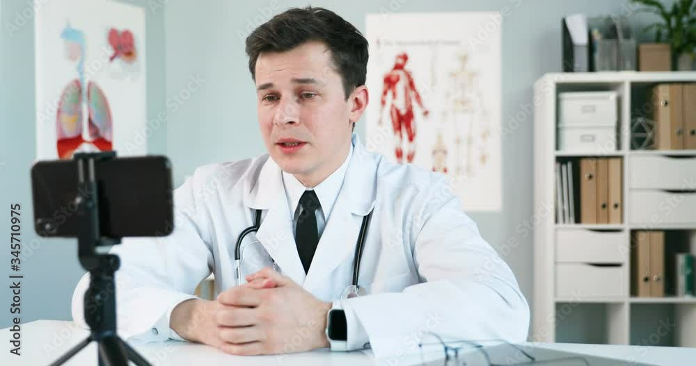 Male Caucasian physician in white gown sitting at table in cabinet. Doctor videochatting on mobile phone and consulting patient via webcam. Medic having videochat and consultation online on smartphone Male Caucasian physician in white gown sitting at table in cabinet. Doctor videochatting on mobile phone and consulting patient via webcam. Medic having videochat and consultation online on smartphone