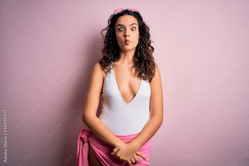 Beautiful woman with curly hair on vacation wearing white swimsuit over pink background making fish face with lips, crazy and comical gesture. Funny expression.