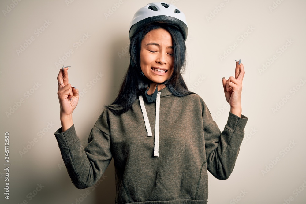 Young beautiful chinese woman wearing bike helmet over isolated white background gesturing finger crossed smiling with hope and eyes closed. Luck and superstitious concept. Young beautiful chinese woman wearing bike helmet over isolated white background gesturing finger crossed smiling with hope and eyes closed. Luck and superstitious concept.