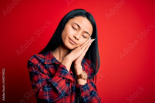 Young beautiful chinese woman wearing casual shirt over isolated red background sleeping tired dreaming and posing with hands together while smiling with closed eyes. © Krakenimages.com