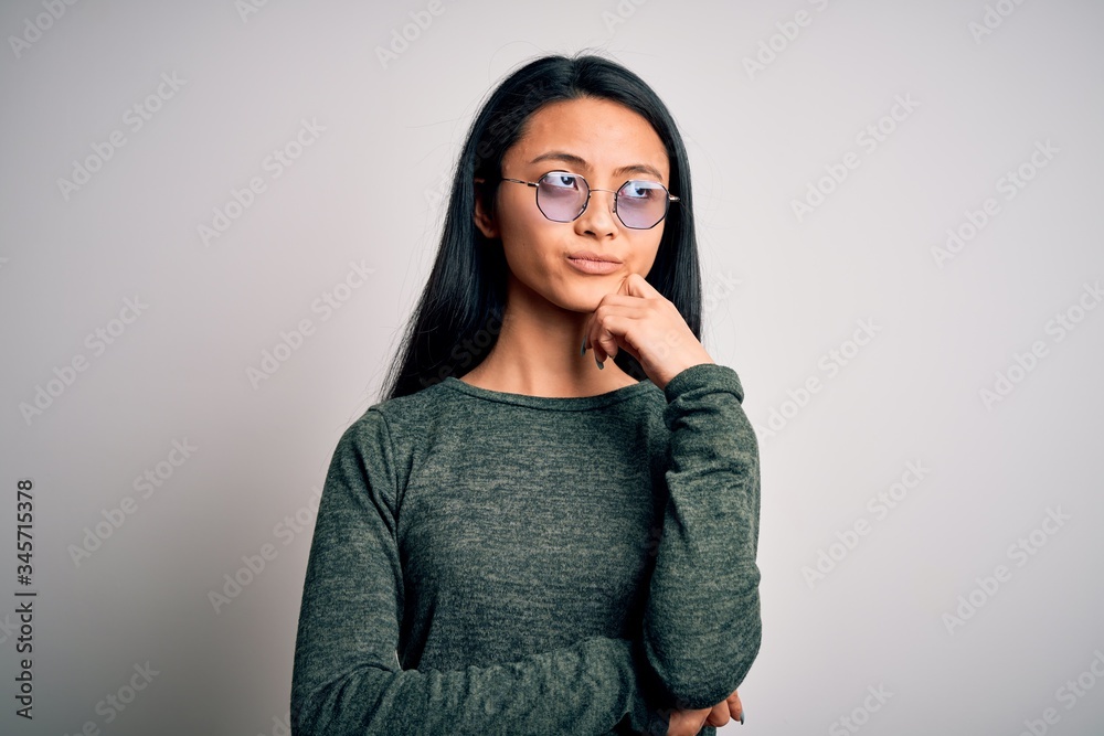 Young beautiful chinese woman wearing glasses and t-shirt over isolated white background with hand on chin thinking about question, pensive expression. Smiling with thoughtful face. Doubt concept. Young beautiful chinese woman wearing glasses and t-shirt over isolated white background with hand on chin thinking about question, pensive expression. Smiling with thoughtful face. Doubt concept.