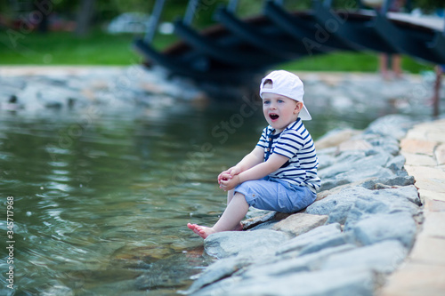 Fototapeta Naklejka Na Ścianę i Meble -  the baby in a white cap sitting by the pond touches cold water with his leg and admires