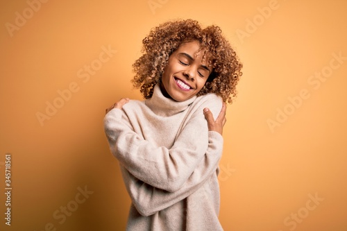 Young beautiful african american woman wearing turtleneck sweater over yellow background Hugging oneself happy and positive, smiling confident. Self love and self care © Krakenimages.com