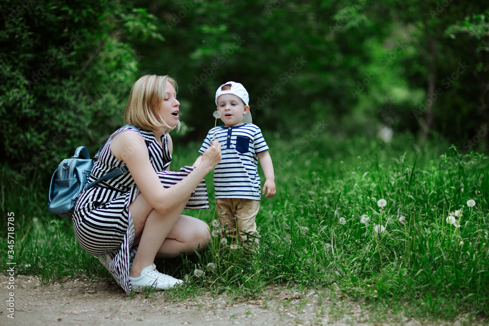 Fototapeta premium young beautiful mother with a small son, frozen from admiration and anticipation, opening his mouth to blow on a flowering dandelion in a green juicy meadow