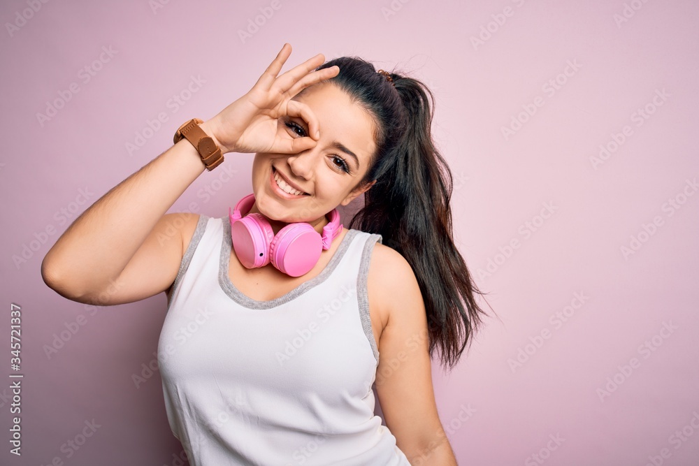 Young brunette fitness woman wearing gym headphones over pink isolated background doing ok gesture with hand smiling, eye looking through fingers with happy face. Young brunette fitness woman wearing gym headphones over pink isolated background doing ok gesture with hand smiling, eye looking through fingers with happy face.