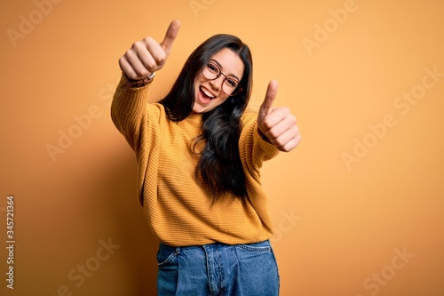 Young brunette woman wearing glasses and casual sweater over yellow isolated background approving doing positive gesture with hand, thumbs up smiling and happy for success. Winner gesture. photo
