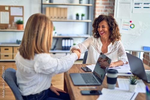 Two middle age beautiful businesswomen smiling happy and confident working together. Shaking hands with smile on face for agreement at the office
