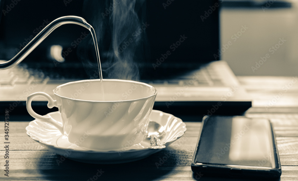 A white cup of coffee with hot smoke in stainless pot, Morning coffee time with notebook computer and work equipment, Normal practice by working at home during covid-19 outbreak around the world. A white cup of coffee with hot smoke in stainless pot, Morning coffee time with notebook computer and work equipment, Normal practice by working at home during covid-19 outbreak around the world.