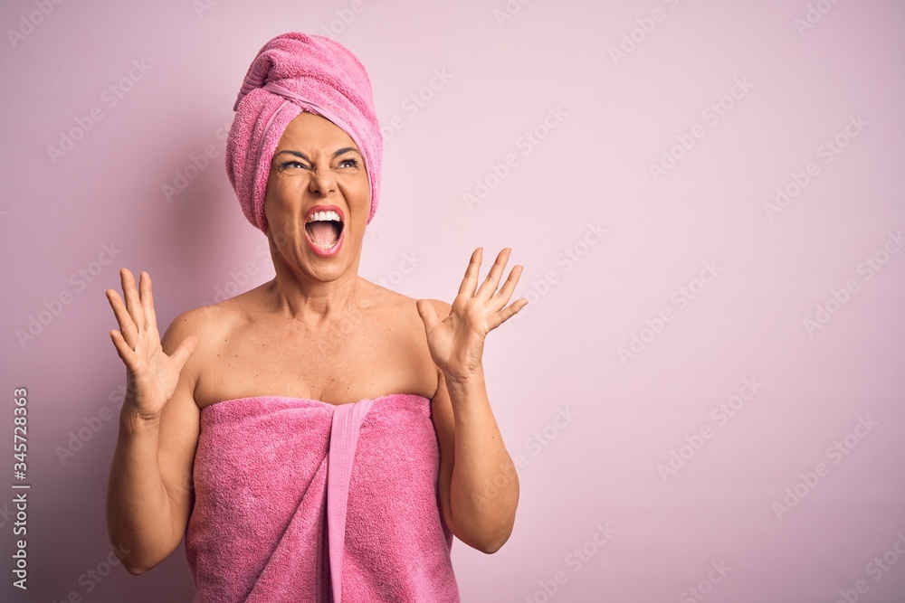 Middle age woman wearing bath towel from beauty body care over pink background crazy and mad shouting and yelling with aggressive expression and arms raised. Frustration concept. Middle age woman wearing bath towel from beauty body care over pink background crazy and mad shouting and yelling with aggressive expression and arms raised. Frustration concept.