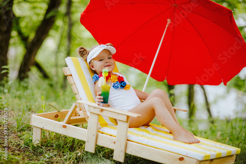 Funny little girl in white swimsuit with flowers relaxing on the striped yellow-and-white deck chair lounger with red sun umbrella