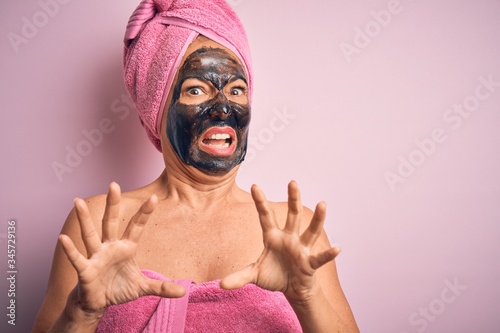Middle age brunette woman wearing beauty black face mask over isolated pink background afraid and terrified with fear expression stop gesture with hands, shouting in shock. Panic concept.