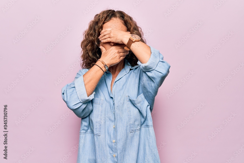 Middle age beautiful woman wearing casual denim shirt standing over pink background Covering eyes and mouth with hands, surprised and shocked. Hiding emotion Middle age beautiful woman wearing casual denim shirt standing over pink background Covering eyes and mouth with hands, surprised and shocked. Hiding emotion