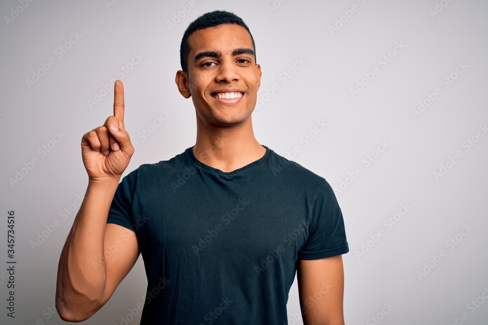 Young handsome african american man wearing casual t-shirt standing over white background showing and pointing up with finger number one while smiling confident and happy. Young handsome african american man wearing casual t-shirt standing over white background showing and pointing up with finger number one while smiling confident and happy.