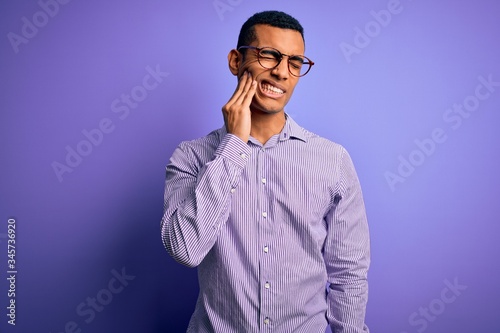 Handsome african american man wearing striped shirt and glasses over purple background touching mouth with hand with painful expression because of toothache or dental illness on teeth. Dentist