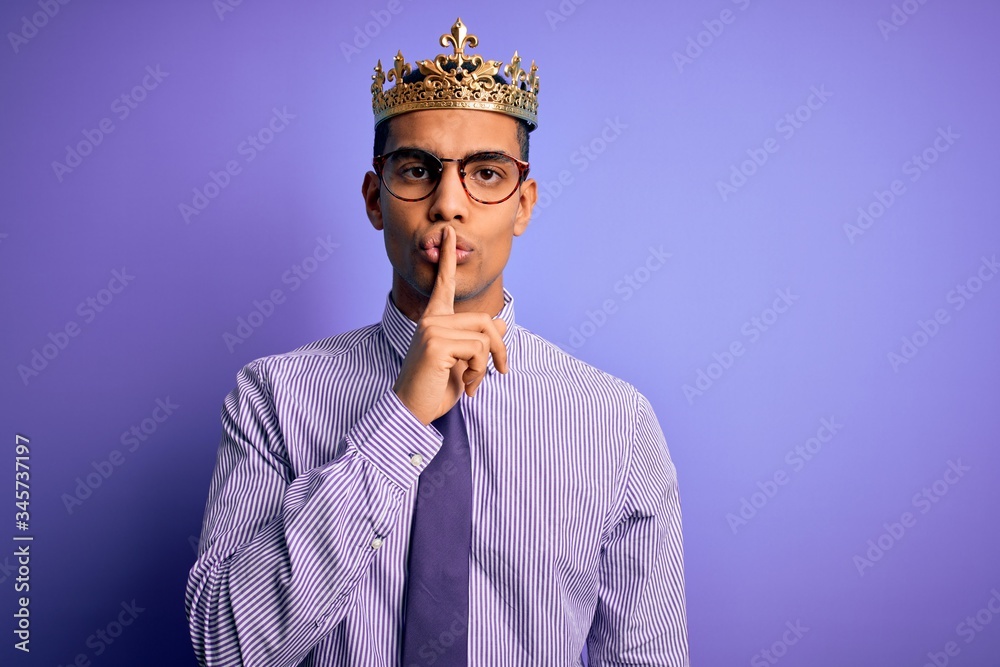 Young handsome african american man wearing golden crown of king over purple background asking to be quiet with finger on lips. Silence and secret concept. Young handsome african american man wearing golden crown of king over purple background asking to be quiet with finger on lips. Silence and secret concept.