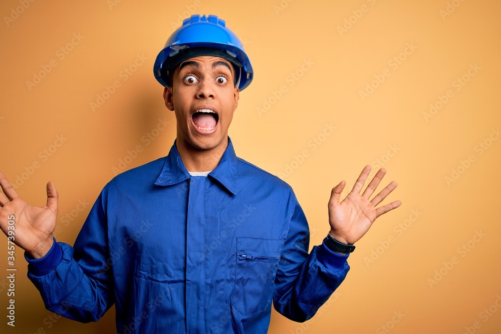 Young handsome african american worker man wearing blue uniform and security helmet celebrating crazy and amazed for success with arms raised and open eyes screaming excited. Winner concept