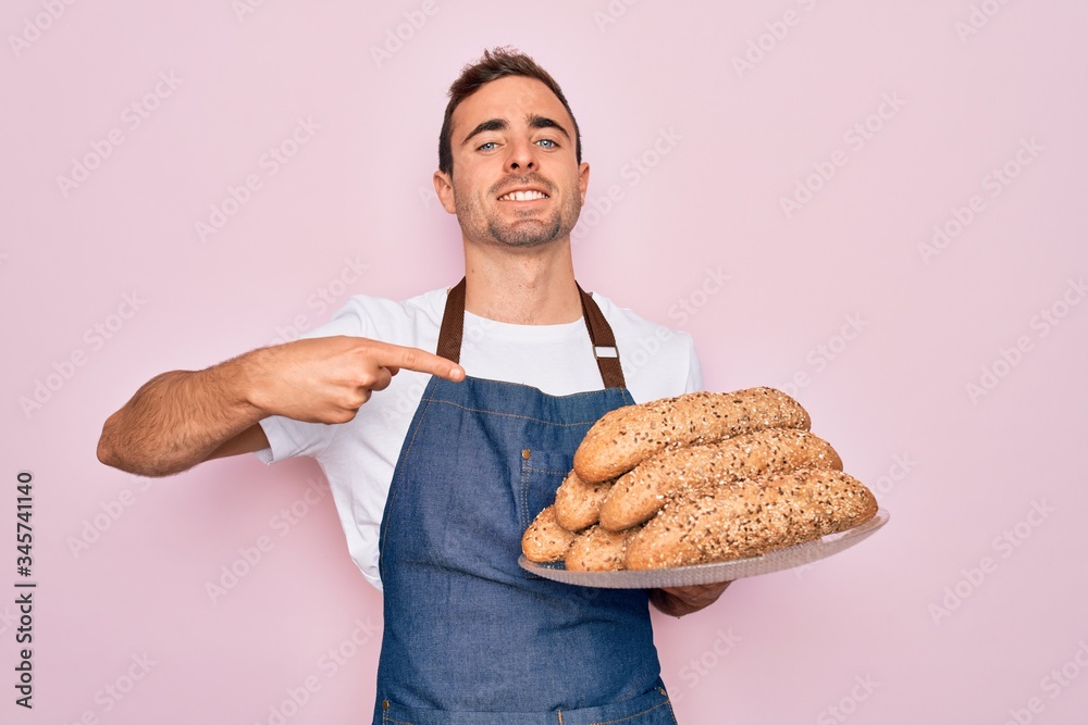 Young handsome baker man with blue eyes wearing apron holding tray with wholmeal bread very happy pointing with hand and finger Young handsome baker man with blue eyes wearing apron holding tray with wholmeal bread very happy pointing with hand and finger