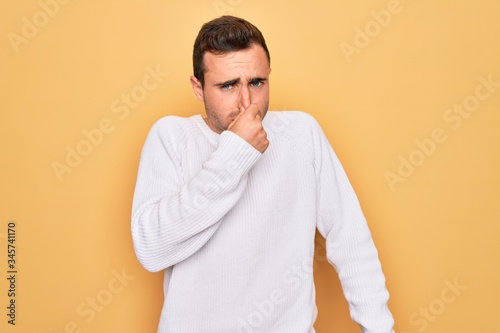 Young handsome man with blue eyes wearing casual sweater standing over yellow background smelling something stinky and disgusting, intolerable smell, holding breath with fingers on nose. Bad smell