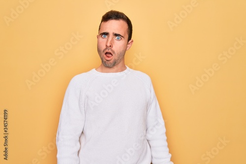 Young handsome man with blue eyes wearing casual sweater standing over yellow background In shock face, looking skeptical and sarcastic, surprised with open mouth