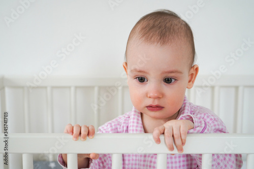 A small child in pink pajamas is standing in the crib. The kid woke up and waits for his parents to pull him out of the crib. Baby starts to cry