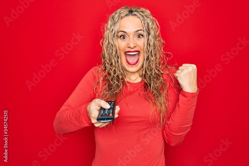 Young beautiful blonde woman using television remote control over isolated red background screaming proud, celebrating victory and success very excited with raised arms