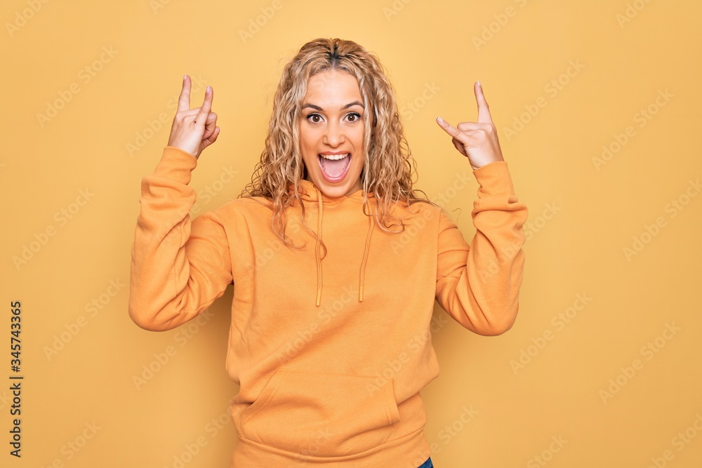 Obraz premium Young beautiful blonde sporty woman wearing casual sweatshirt over yellow background shouting with crazy expression doing rock symbol with hands up. Music star. Heavy music concept.