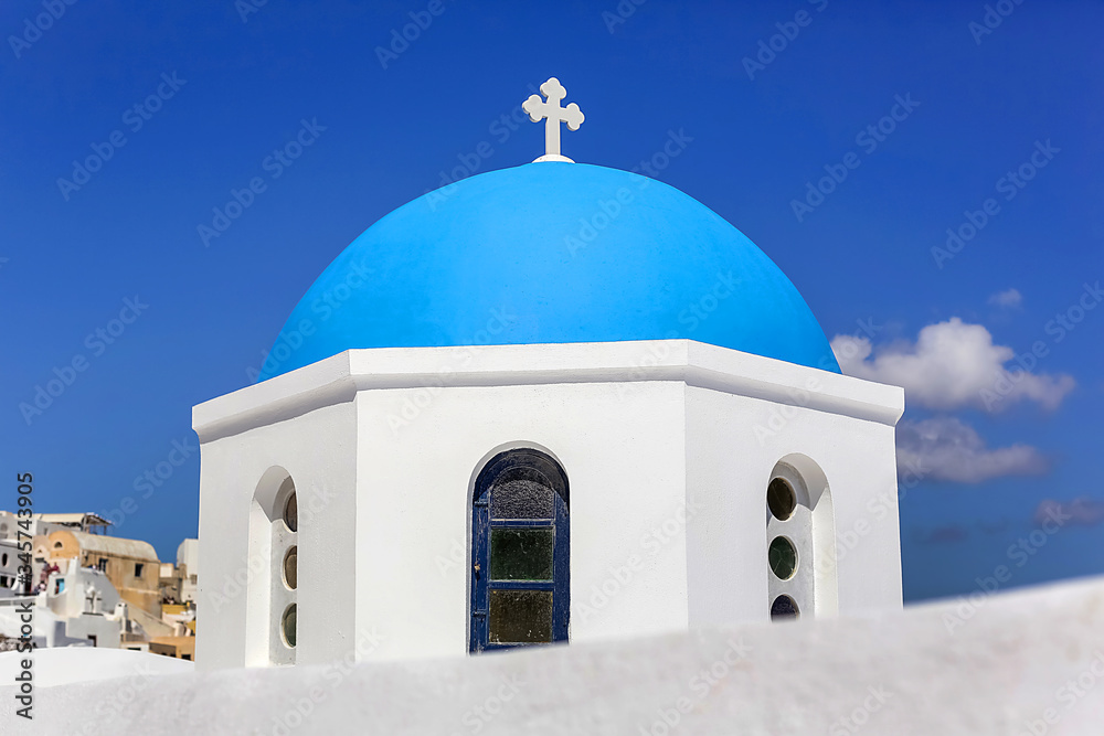 Close-up of the round dome of the Church with a blue cross, white stone walls against the blue sky in Greece on Santorini