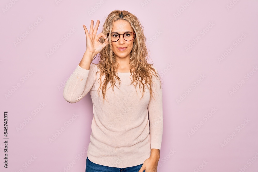 Young beautiful blonde woman wearing casual sweater and glasses over pink background smiling positive doing ok sign with hand and fingers. Successful expression. Young beautiful blonde woman wearing casual sweater and glasses over pink background smiling positive doing ok sign with hand and fingers. Successful expression.