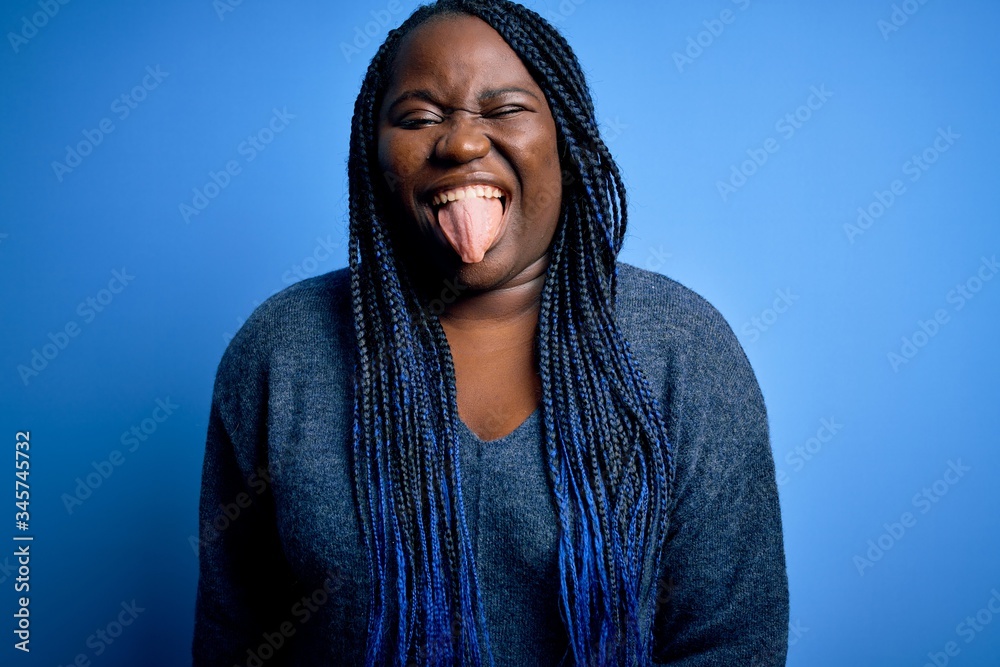 African american plus size woman with braids wearing casual sweater over blue background sticking tongue out happy with funny expression. Emotion concept. African american plus size woman with braids wearing casual sweater over blue background sticking tongue out happy with funny expression. Emotion concept.
