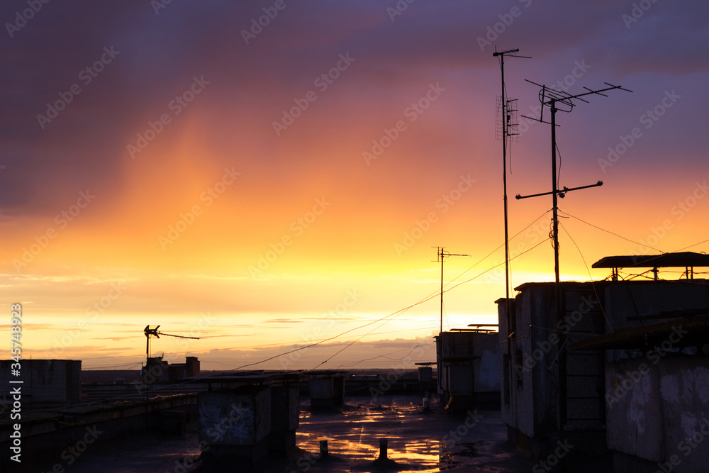 Sunset on the roof of a multi-storey building overlooking the yellow-blue sky and antennas. Roof after the rain