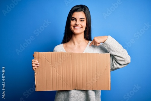 Young beautiful brunette activist woman holding blank cardboard banner protesting with surprise face pointing finger to himself