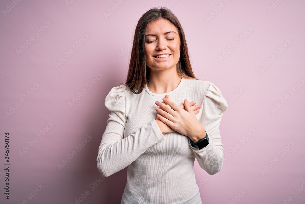 Young beautiful woman with blue eyes wearing casual white t-shirt over pink background smiling with hands on chest with closed eyes and grateful gesture on face. Health concept. Young beautiful woman with blue eyes wearing casual white t-shirt over pink background smiling with hands on chest with closed eyes and grateful gesture on face. Health concept.
