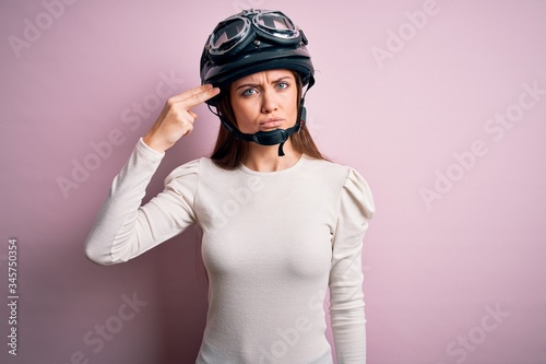 Young beautiful motorcyclist woman with blue eyes wearing moto helmet over pink background Shooting and killing oneself pointing hand and fingers to head like gun, suicide gesture.