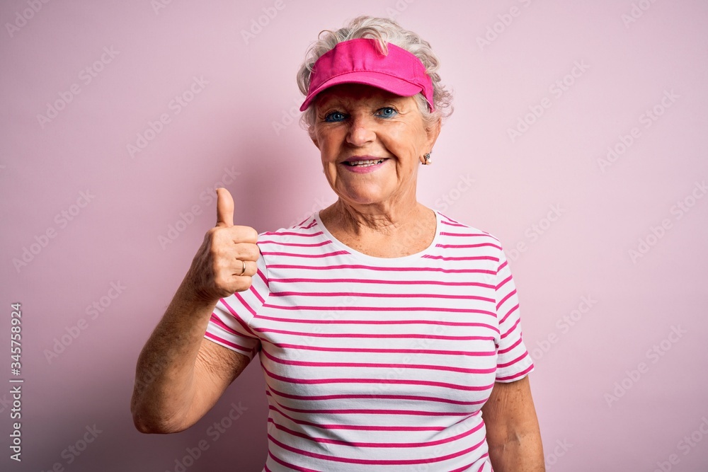 Senior beautiful sporty woman wearing sport cap standing over isolated pink background doing happy thumbs up gesture with hand. Approving expression looking at the camera showing success. Senior beautiful sporty woman wearing sport cap standing over isolated pink background doing happy thumbs up gesture with hand. Approving expression looking at the camera showing success.