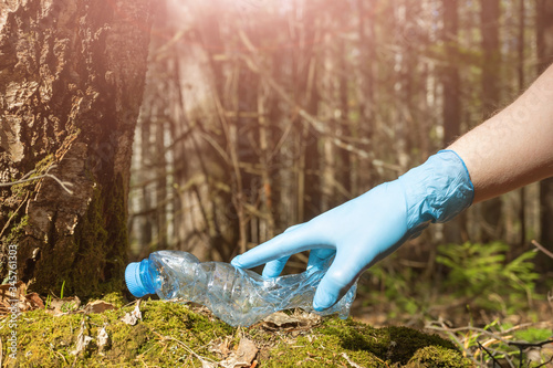 An empty crumpled plastic bottle is lying on the ground in the forest. Hand in a blue rubber glove. The concept of garbage, forest cleaning, negative impact on nature, ecology, environment, cleanup. photo