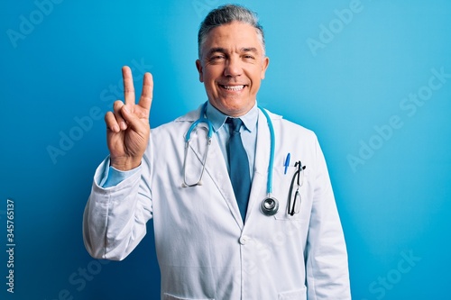 Middle age handsome grey-haired doctor man wearing coat and blue stethoscope showing and pointing up with fingers number two while smiling confident and happy.