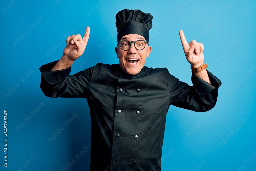 Middle age handsome grey-haired chef man wearing cooker uniform and hat smiling amazed and surprised and pointing up with fingers and raised arms. Middle age handsome grey-haired chef man wearing cooker uniform and hat smiling amazed and surprised and pointing up with fingers and raised arms.