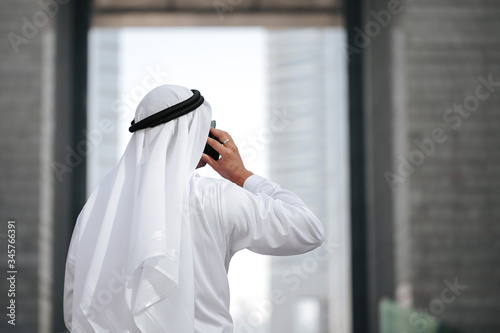 Emirati wearing traditional kandura talking with smart phone. Arab guy in gulf country touring around the urban city lifestyle. photo