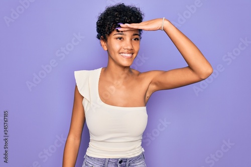Young beautiful african american afro woman wearing casual t-shirt over purple background very happy and smiling looking far away with hand over head. Searching concept.