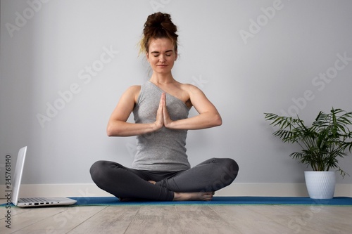 A girl in gray t-shirt and leggings sit in yoga position at home on blue rug near palm tree in white pot and white laptop on ceramic wooden tile