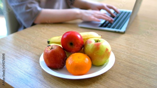 A plate of fruits on a table with the hands of a woman on a laptop in background