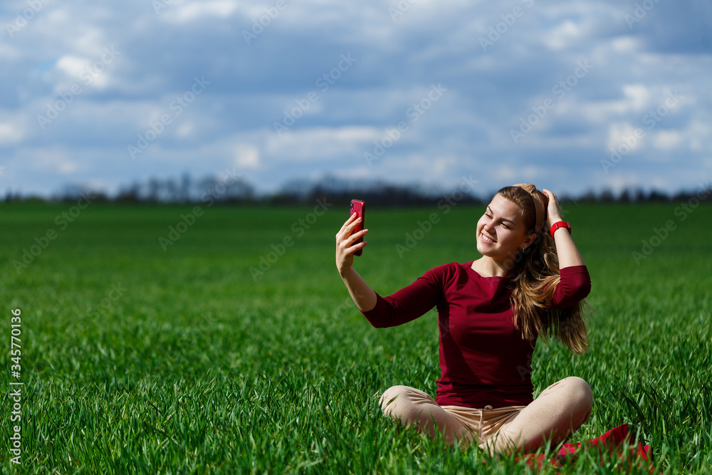 Young beautiful woman student with a phone in her hands sitting on the grass. Girl takes selfies and takes selfie pictures. She smiles and enjoys a warm day. Concept photo on smartphone Young beautiful woman student with a phone in her hands sitting on the grass. Girl takes selfies and takes selfie pictures. She smiles and enjoys a warm day. Concept photo on smartphone
