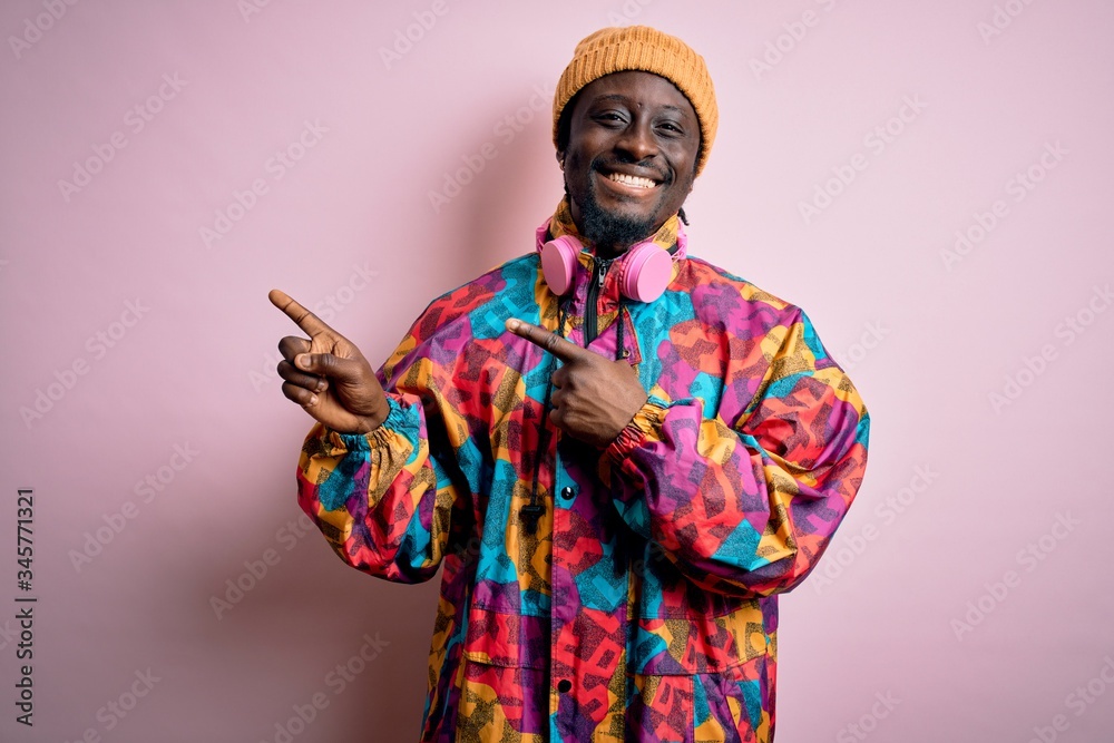 Young handsome african american man wearing colorful coat and cap over pink background smiling and looking at the camera pointing with two hands and fingers to the side.