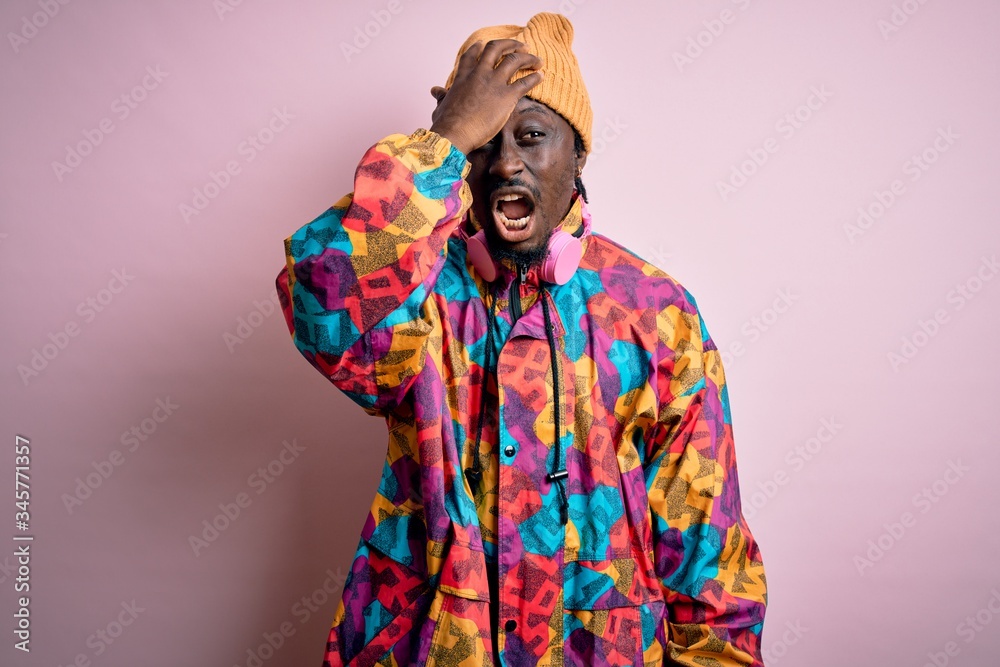 Young handsome african american man wearing colorful coat and cap over pink background surprised with hand on head for mistake, remember error. Forgot, bad memory concept.