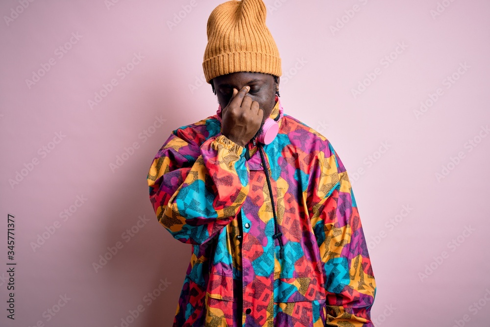 Young handsome african american man wearing colorful coat and cap over pink background tired rubbing nose and eyes feeling fatigue and headache. Stress and frustration concept.